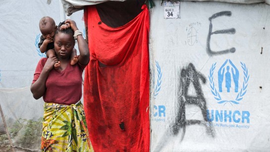 En mamma bär sitt barn på axlarna utan för ett tält från UNHCR.