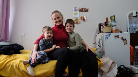 En kvinna och hennes två barn sitter på en säng. De tittar in i kameran och ler.