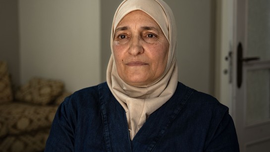 Shaza, en 59-årig kvinna från Syrien, står i ett vardagsrum och tittar framför sig med orolig blick. Hon bär en ljus sjal och mörkblå kläder.